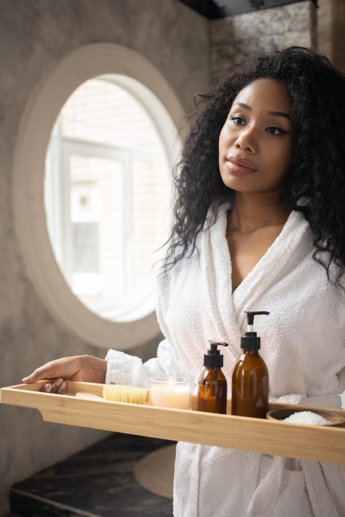 Young ethnic female in white bathrobe holding wooden tray with skin care products