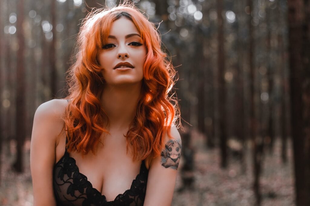 Sensual female with red hair and in lace bra standing in autumn woods and looking at camera