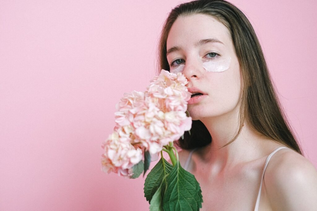 Charming young female model with long hair touching face with fresh hydrangea while standing with facial mask on pink background